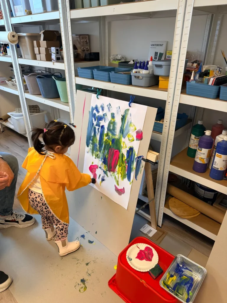 Een peuter in een geel schortje schildert met felle kleuren in het atelier